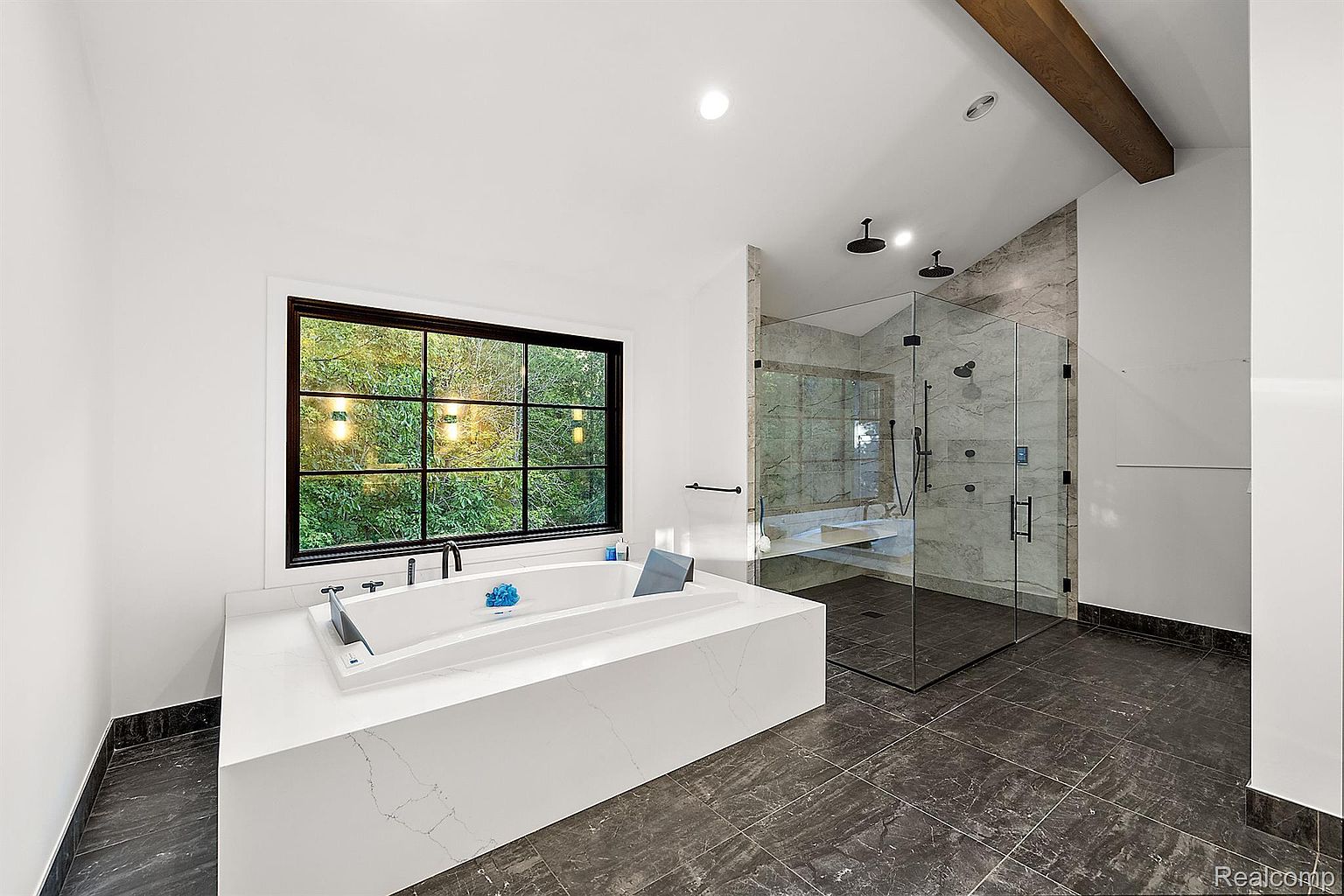 This is a luxurious primary bathroom featuring a modern freestanding bathtub with a sleek faucet, positioned beneath a large window offering a view of lush greenery. A glass-enclosed shower with dual showerheads and marble-like tiling adds to the spa-like ambiance. The dark gray tiled floor provides a sophisticated contrast to the white walls and fixtures, creating a serene and upscale retreat.