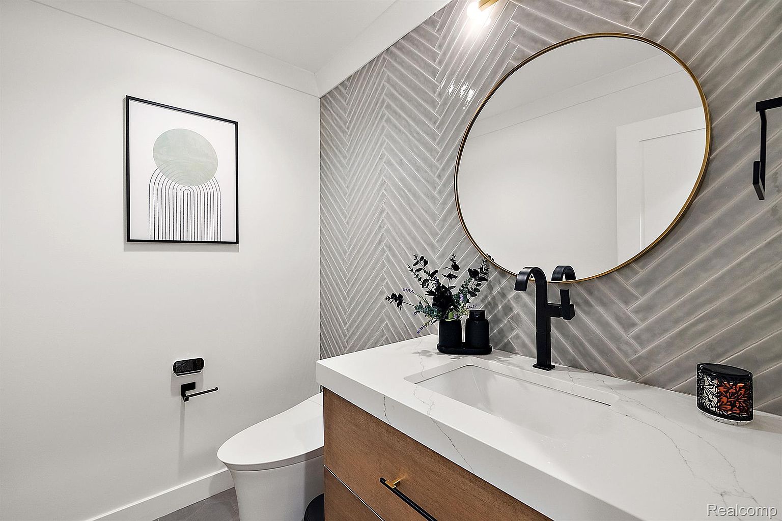 This is a stylish guest bathroom featuring a modern design. The vanity has a white countertop and wood-look cabinetry, complemented by a black faucet and a round mirror with a gold frame. The wall behind the vanity is tiled in a herringbone pattern, adding texture and visual interest. A framed art piece hangs on the adjacent wall, and a toilet with a black toilet paper holder is visible.