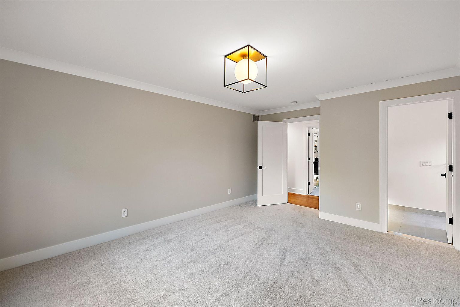 This is a well-lit bedroom featuring neutral-toned walls and light gray carpeting. The room has white trim and baseboards, adding a clean and modern touch. A contemporary light fixture hangs from the ceiling, and two doorways lead to other areas of the house, creating an open and airy feel.