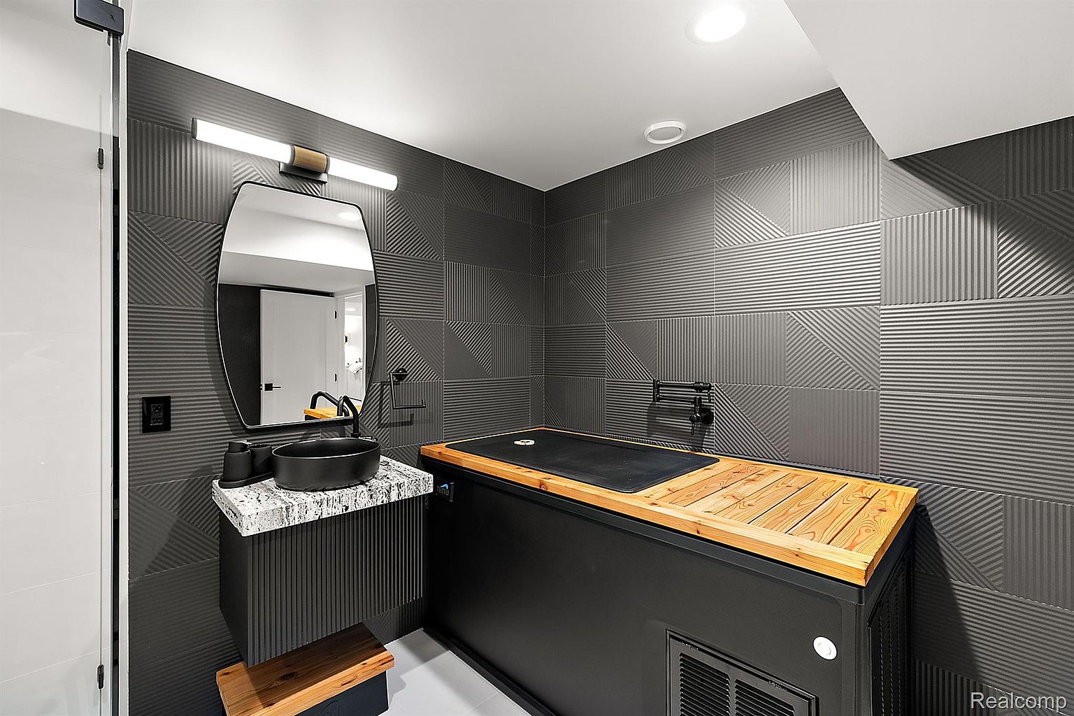 This modern bathroom features a unique design with textured gray tile walls and a black vessel sink on a granite countertop. A large, dark-toned structure with a wooden top dominates the space, possibly a custom-built feature. The room is well-lit with a contemporary light fixture above the mirror, creating a sophisticated and stylish atmosphere.