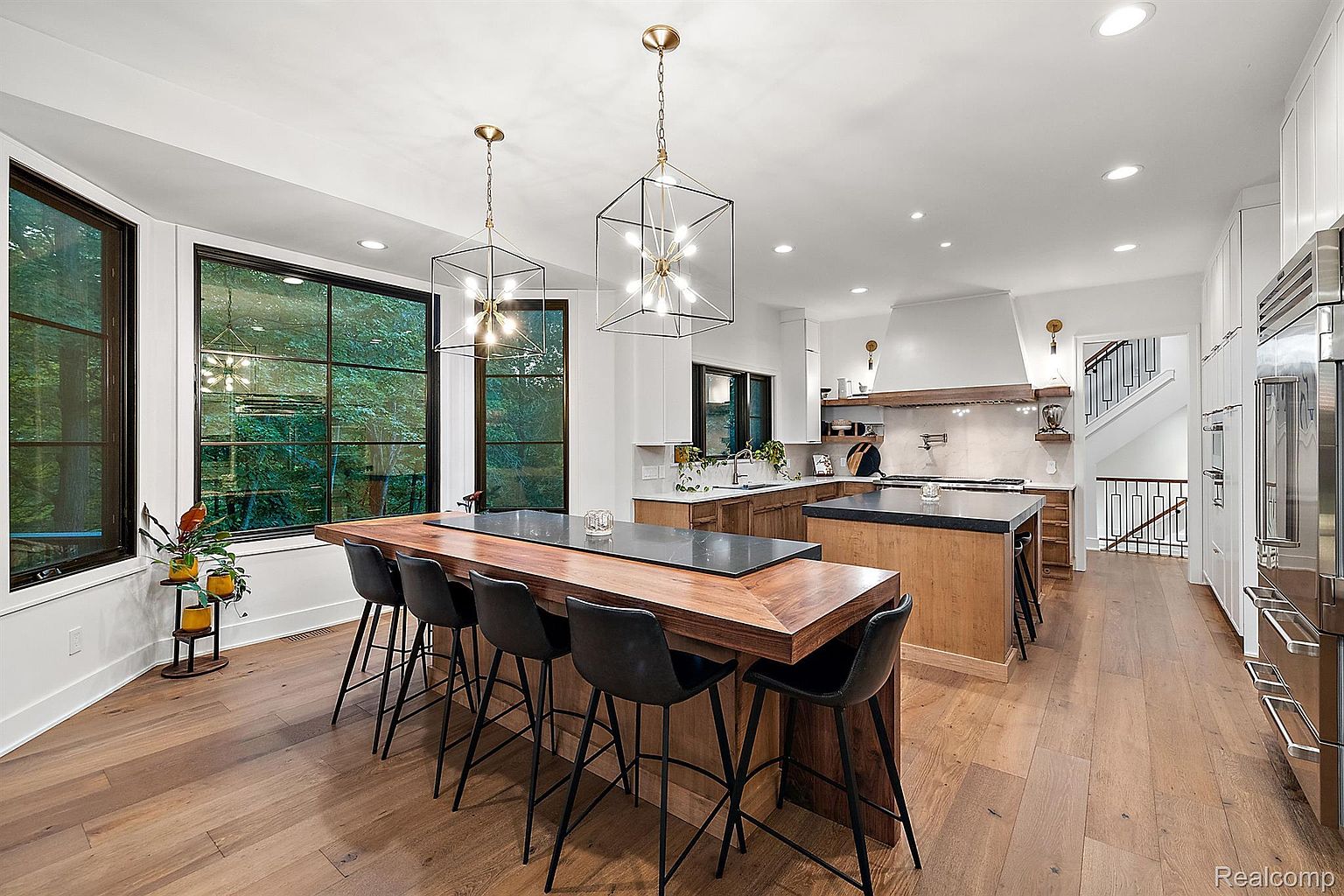 This is a spacious, modern kitchen featuring a large island with seating, custom cabinetry, and high-end appliances. The kitchen boasts a blend of wood and dark countertops, complemented by contemporary pendant lighting and large windows that provide ample natural light. The open layout and hardwood floors contribute to a warm and inviting atmosphere.