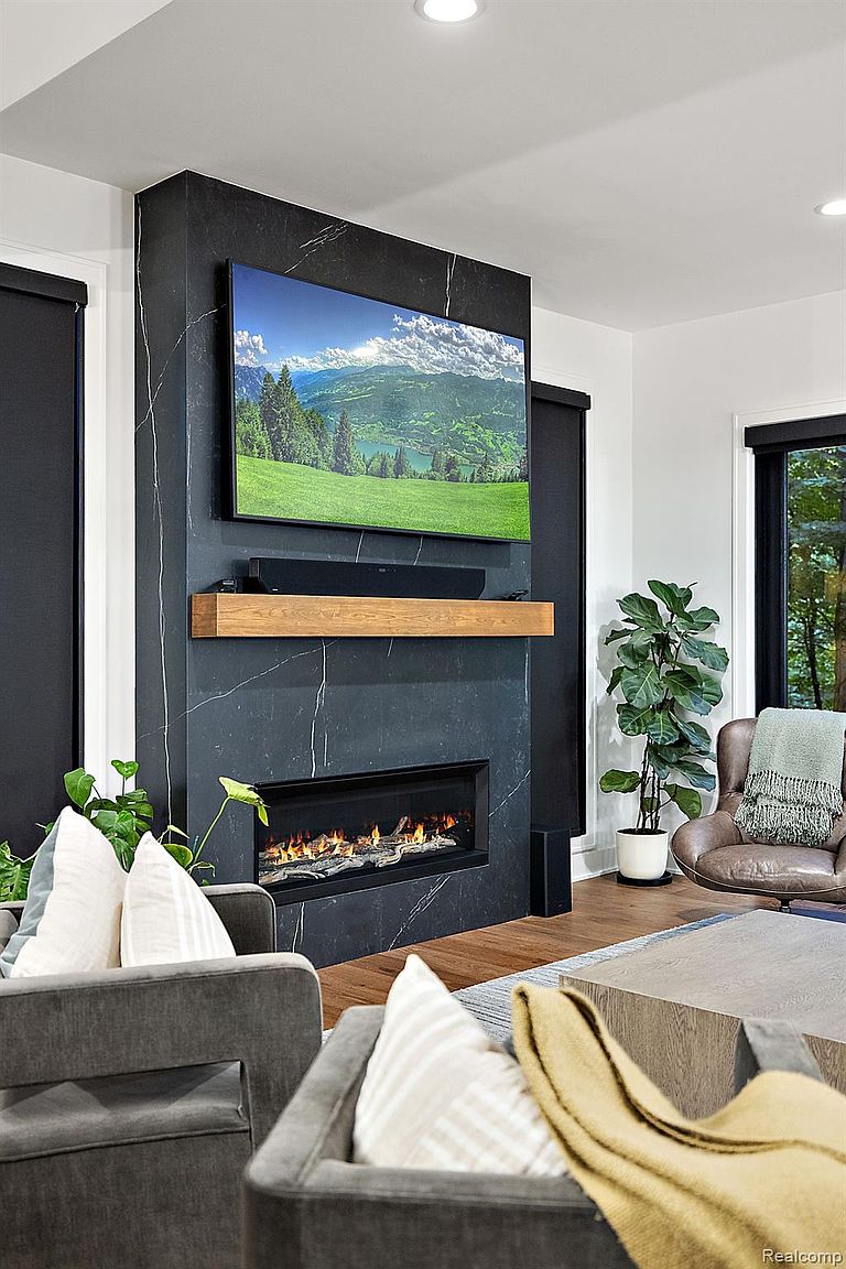 The image showcases a modern living room with a striking black marble fireplace as a focal point, above which hangs a large television displaying a scenic landscape. The room is furnished with contemporary gray armchairs and a neutral-toned coffee table, complemented by a tall potted plant adding a touch of nature. The overall aesthetic is sophisticated and inviting, emphasizing clean lines and a comfortable atmosphere.