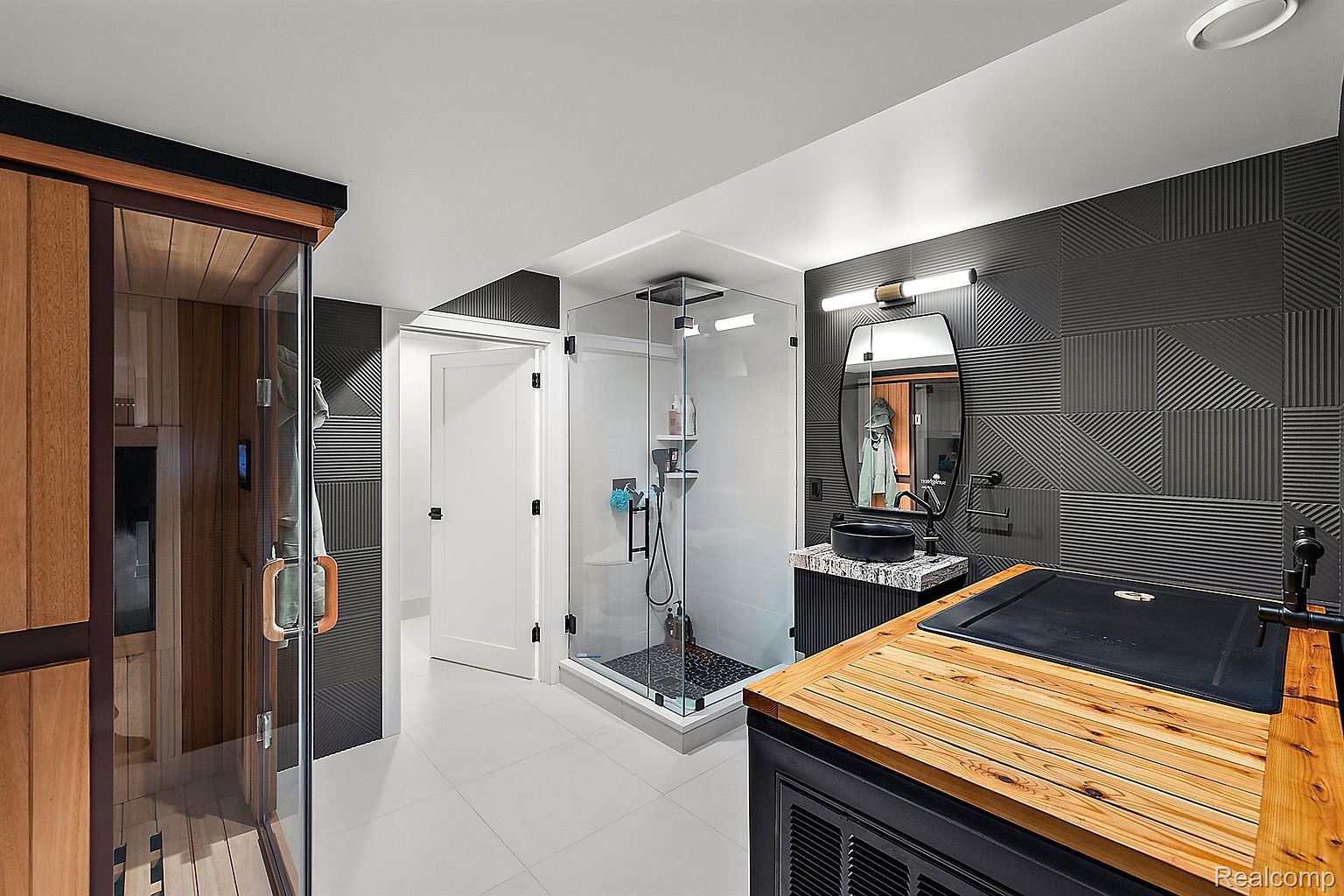 This is a modern primary bathroom featuring a sauna, a glass-enclosed shower with black hardware, and a sleek vanity with a black vessel sink. The walls are adorned with textured gray tiles, and the floor is covered in large format light gray tiles. The overall design is contemporary and luxurious, creating a spa-like atmosphere.