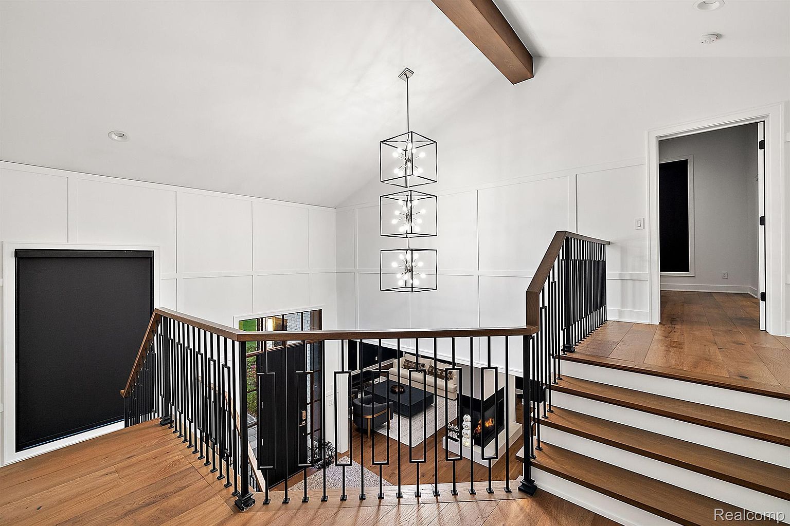This interior shot showcases a modern hallway and staircase. The staircase features wooden steps with white risers and a black metal railing with a wooden handrail. A striking, multi-tiered cube chandelier hangs above, illuminating the space and adding a touch of contemporary elegance.