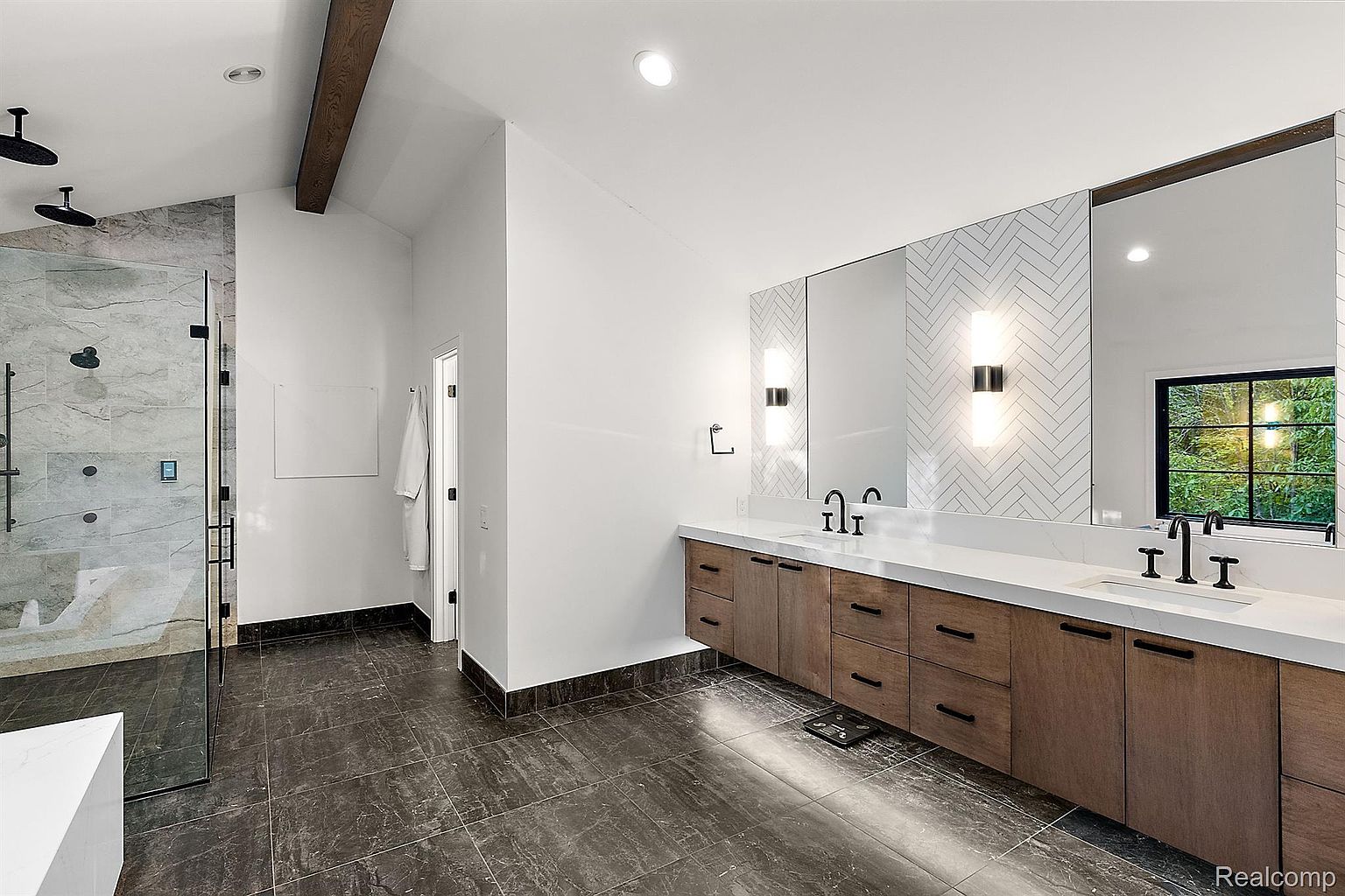 This is a primary bathroom featuring a double vanity with wood cabinets and a white countertop. The mirrors are framed and flanked by sconces, with a herringbone tile backsplash adding visual interest. A glass-enclosed shower with marble-like tile is visible to the left, and dark tile flooring runs throughout the space, creating a modern and luxurious feel.