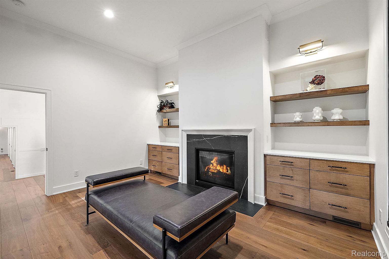 This is an interior shot of a modern living room featuring a fireplace with a black marble surround and a black firebox. The room is decorated with built-in shelving units on either side of the fireplace, and a black leather bench sits in the center of the room. The hardwood floors add warmth to the space, and the white walls create a bright and airy atmosphere.