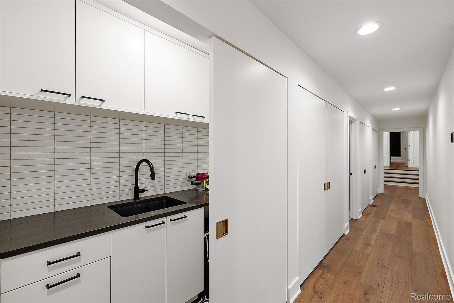 This interior shot showcases a modern hallway with a glimpse of a laundry area. The hallway features sleek, white walls and doors, complemented by warm-toned hardwood flooring. Recessed lighting illuminates the space, leading towards a staircase and an open doorway, creating a bright and inviting atmosphere.