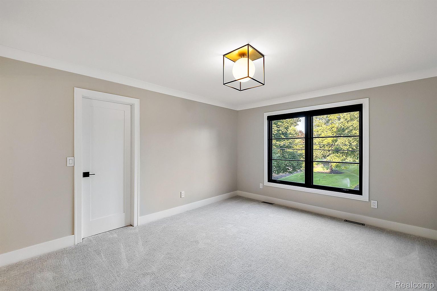 This is a well-lit bedroom featuring neutral-toned walls and light gray carpeting. A modern, geometric light fixture hangs from the ceiling, and a large window provides natural light and a view of the outdoors. A white door with a black handle is visible on the left, contributing to the room's clean and contemporary aesthetic.