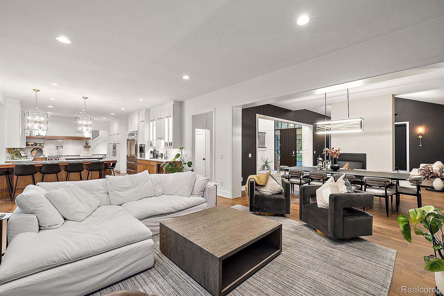 This is an interior shot of a modern living room that seamlessly blends into the dining and kitchen areas. The living room features a large, comfortable white sectional sofa, a dark wood coffee table, and two armchairs, all arranged on a gray area rug. The open floor plan allows for a spacious and airy feel, enhanced by the natural light and the view into the dining area with its modern lighting fixture and fireplace.