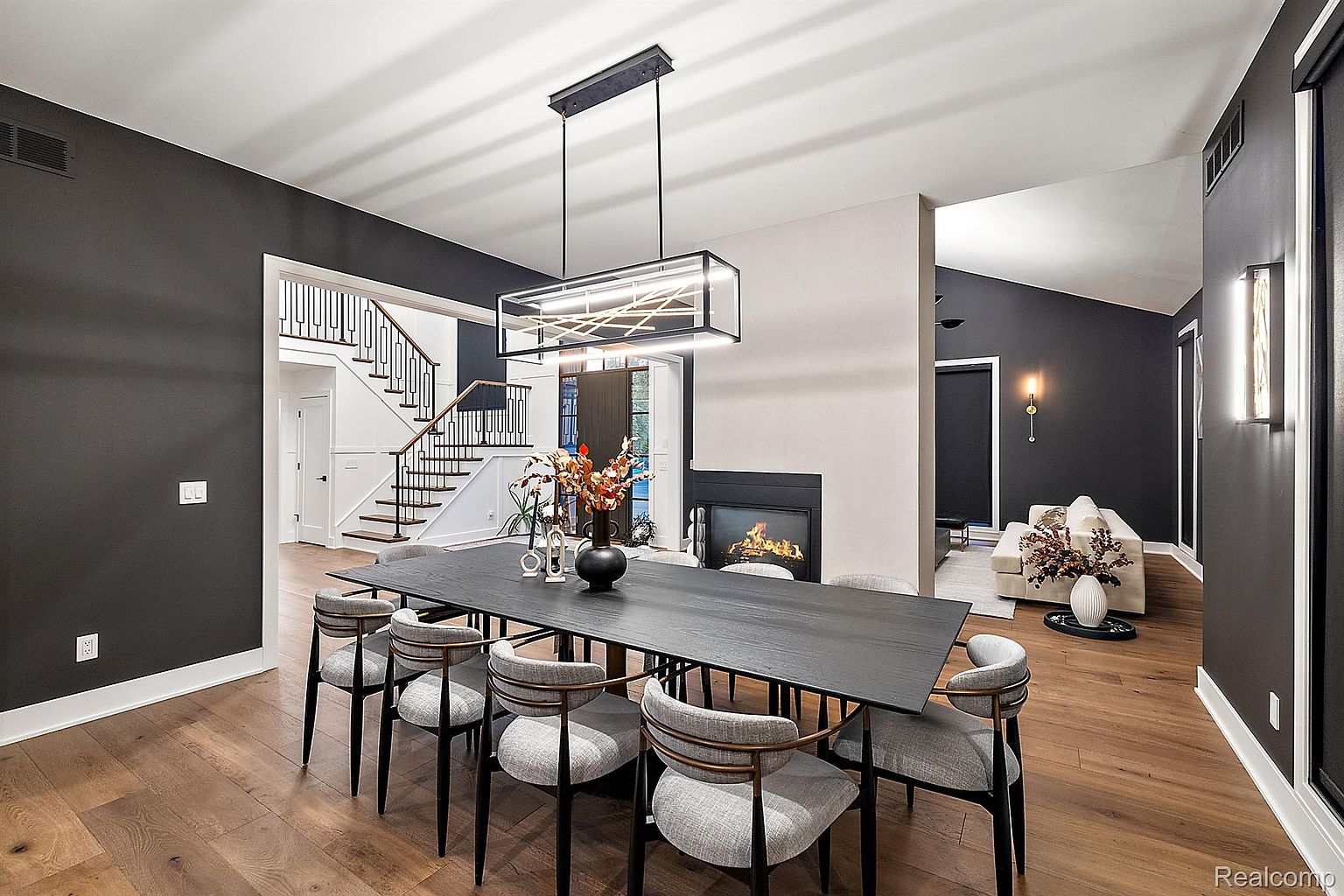 This is an interior shot of a modern dining room featuring a long, dark wood table surrounded by eight chairs with light gray upholstery and black legs. A contemporary rectangular chandelier hangs above the table, and a fireplace is visible in the adjacent living area. The walls are painted in a dark gray, contrasting with the light wood floors, creating a sophisticated and inviting atmosphere.