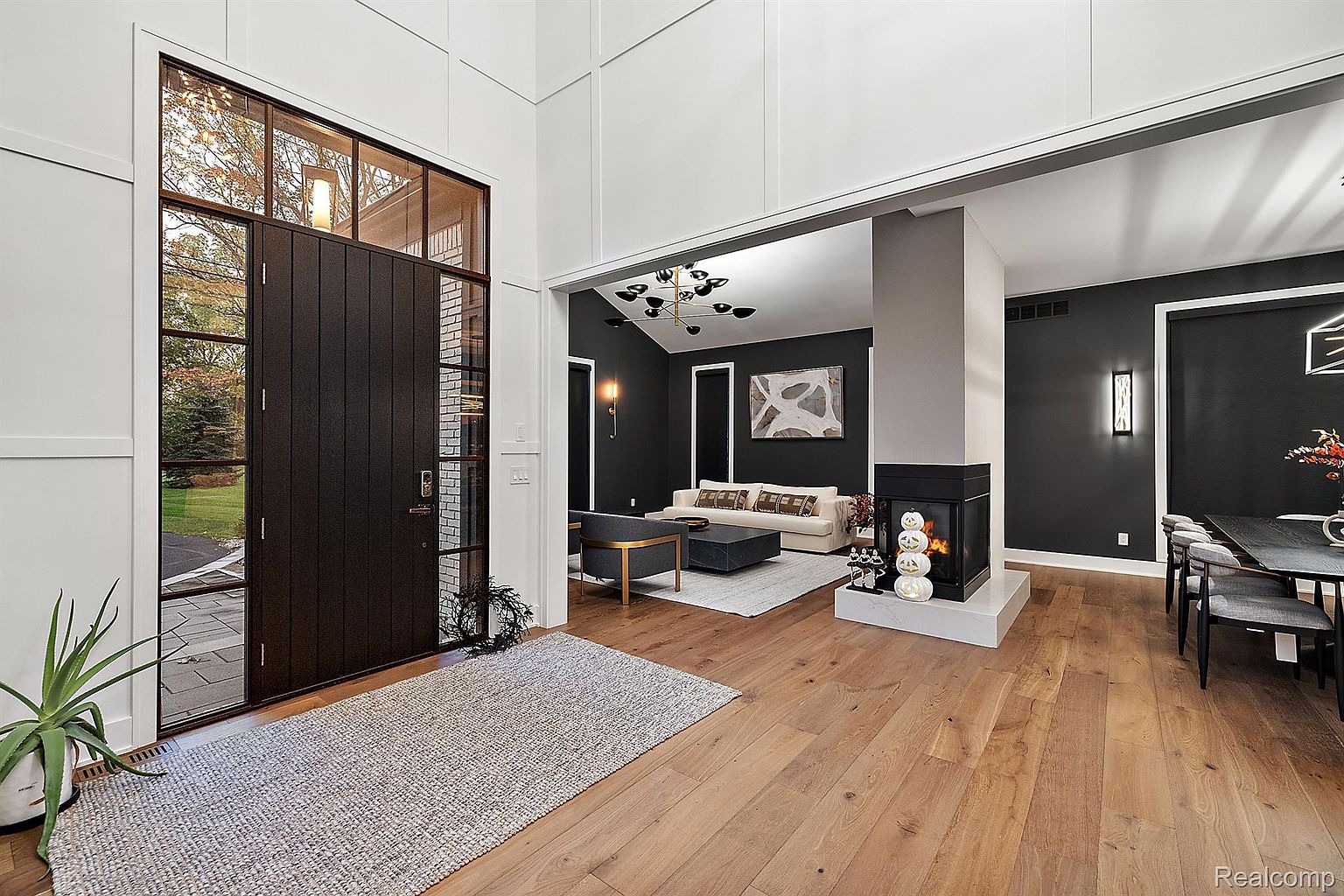 This interior shot showcases a modern living room with dark walls contrasted by white trim and a light wood floor. A fireplace is centrally located, dividing the living room from the dining area. The entryway features a large, dark wood door with glass panels, allowing natural light to flood the space, and a neutral rug adds texture to the floor.
