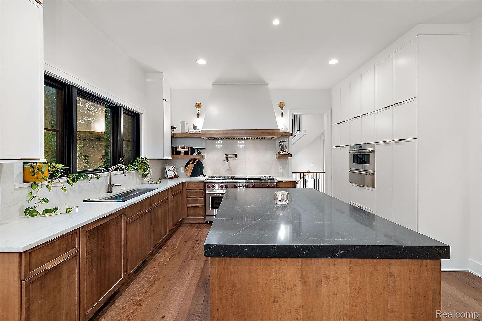 This is a well-lit, modern kitchen featuring a large island with a dark countertop and wooden base. The kitchen includes a stainless steel range with a custom hood, a sink under a window with plants, and white cabinetry with stainless steel accents. The hardwood flooring adds warmth to the space, creating an inviting atmosphere.