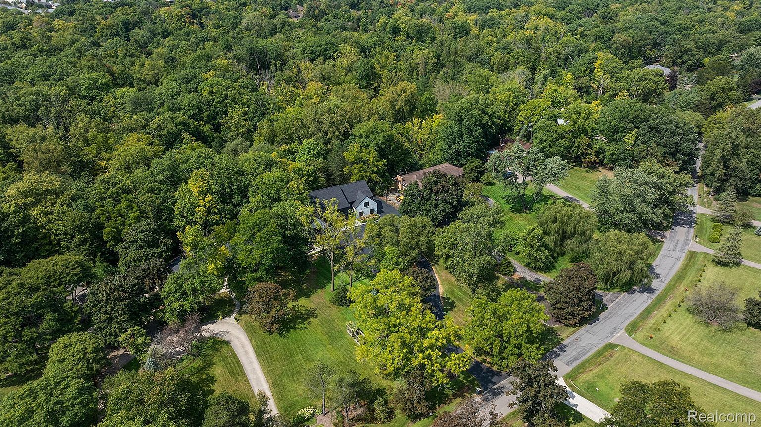 This aerial shot showcases a property nestled within a lush, dense forest. A modern black and white house is visible, surrounded by mature trees and manicured lawns. A winding driveway leads to the house, and a paved road borders the property, suggesting a serene and private setting.
