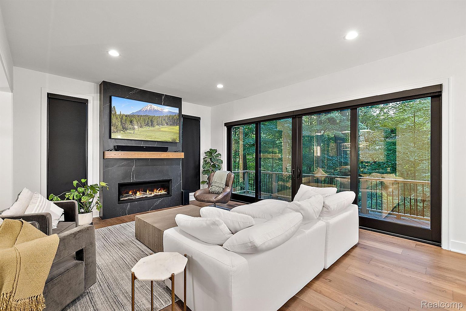 This is a bright and modern living room featuring a sleek fireplace with a mounted television above. A large white sectional sofa faces the fireplace, complemented by a light-colored rug and a stylish side table. Large sliding glass doors provide ample natural light and a view of the outdoor greenery, enhancing the room's open and airy feel.