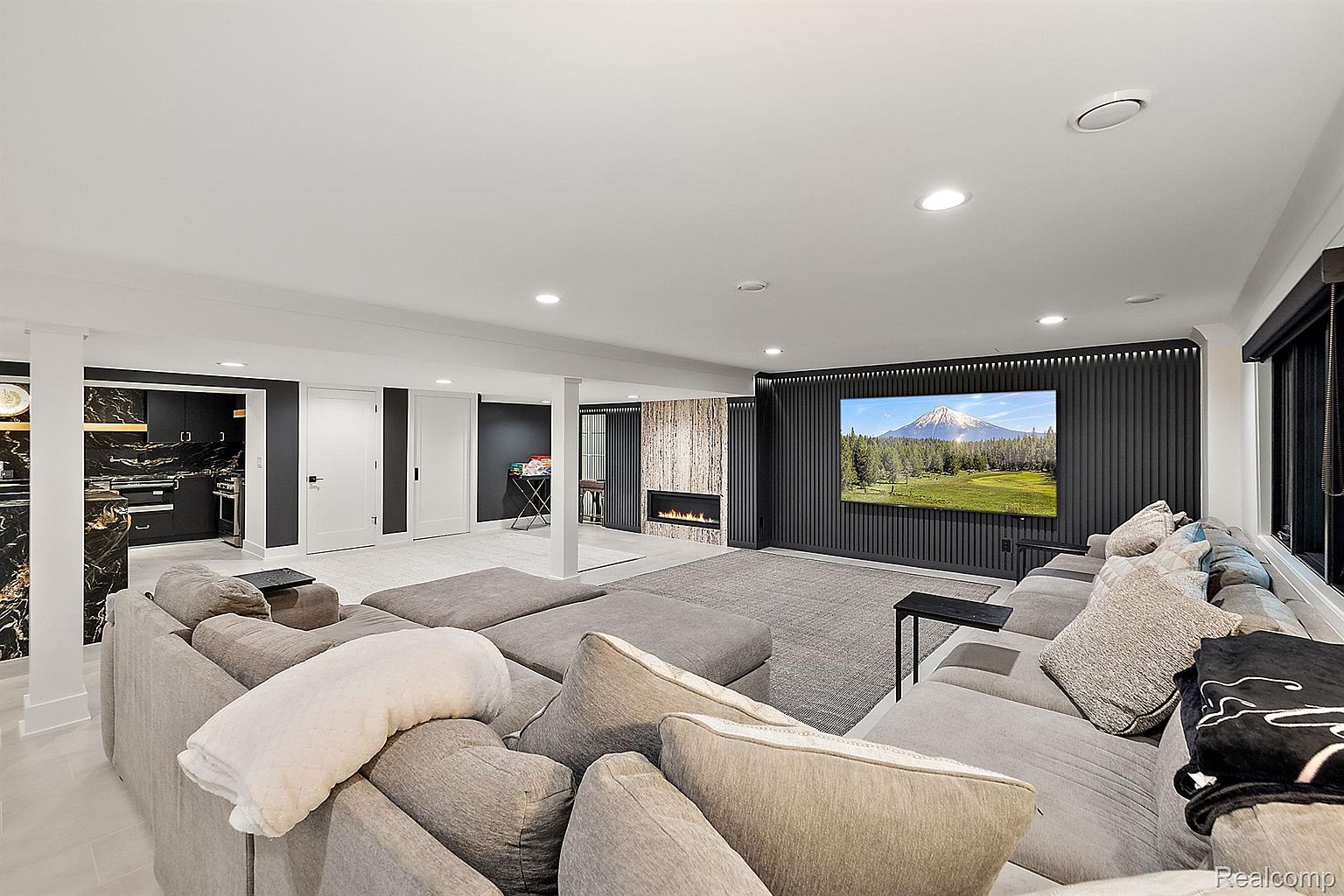 This is a spacious living room featuring a large sectional sofa, a modern fireplace, and a large screen television mounted on a dark paneled wall. The room has a contemporary design with recessed lighting and a neutral color palette, creating a comfortable and inviting atmosphere. The open layout suggests a seamless flow to other areas of the home.