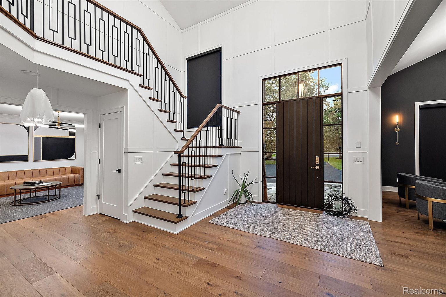This interior shot showcases a grand foyer with hardwood flooring, a staircase with a modern black railing, and a dark wood front door with sidelights. The walls are painted white with wainscoting, creating a bright and inviting space. The overall impression is elegant and contemporary.
