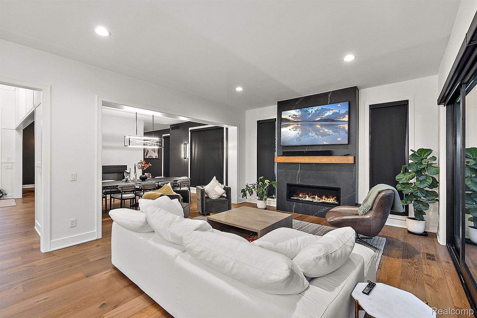 This is an interior shot of a modern living room featuring a white sofa, a dark marble fireplace with a mounted television, and hardwood floors. The room opens to a dining area with a dark accent wall, creating a cohesive and stylish living space. Large windows provide ample natural light, enhancing the room's spacious feel.