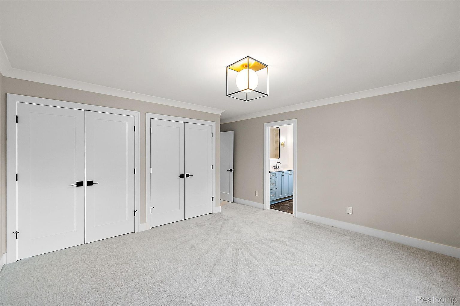The image showcases a bright and airy bedroom with neutral-toned walls and light gray carpeting. Two sets of double doors, painted white with modern black hardware, provide closet space. A contemporary cube-shaped light fixture illuminates the room, and an open doorway reveals a glimpse into an adjacent bathroom with blue cabinetry.