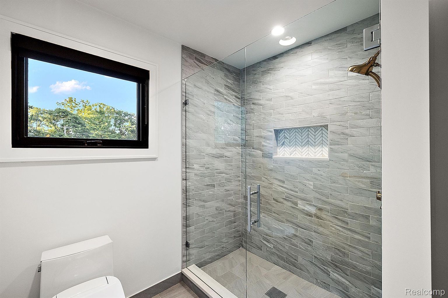 This is a modern bathroom featuring a glass-enclosed shower with gray, horizontally laid tiles and a built-in niche with herringbone pattern tiles. A black-framed window provides natural light, and a toilet is visible in the foreground. The overall impression is clean, contemporary, and well-lit.