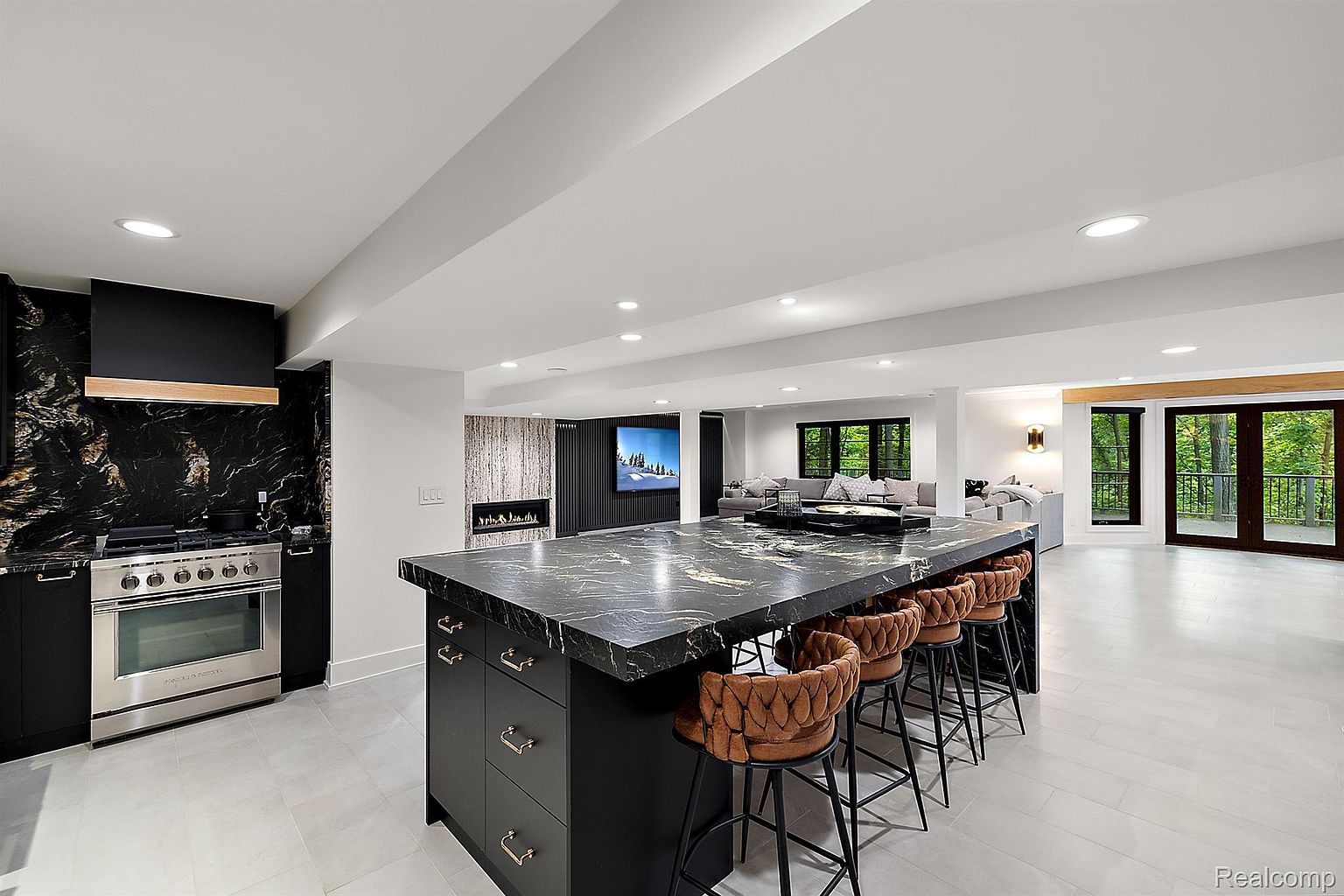 This is a modern, open-concept kitchen featuring a large island with seating, black marble countertops, and stainless steel appliances. The kitchen flows seamlessly into a living area with a fireplace and a view of the outdoors through large windows and doors. The overall impression is luxurious and spacious, perfect for entertaining.