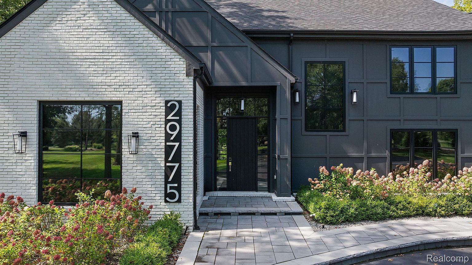 This is a front exterior view of a modern home, showcasing a striking entryway. The house features a combination of white brick and dark gray siding, complemented by black-framed windows and a dark front door. The address number is prominently displayed vertically, and the landscaping includes flowering bushes along the walkway, creating an inviting entrance.