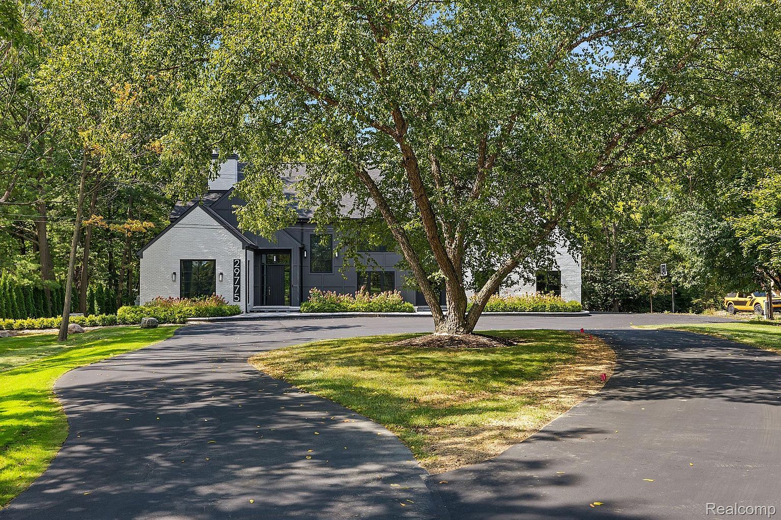 The image showcases the front exterior of a modern home with a striking black and white color scheme. The house features a combination of white brick and dark gray siding, complemented by black-framed windows and a prominent front door. A large, mature tree dominates the front yard, adding a touch of nature and shade to the property, while a circular driveway provides ample parking space.