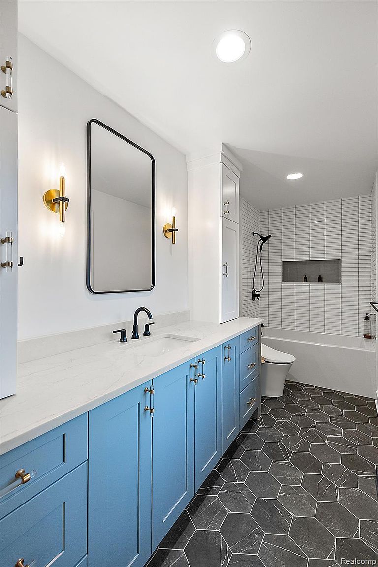 This is a well-lit bathroom featuring a long, light blue vanity with gold hardware and a white countertop. A black-framed mirror hangs above the sink, flanked by gold sconces. The floor is covered in dark, geometric tiles, and a white bathtub and toilet are visible in the background, along with white subway tile in the shower area. The overall impression is modern and stylish.