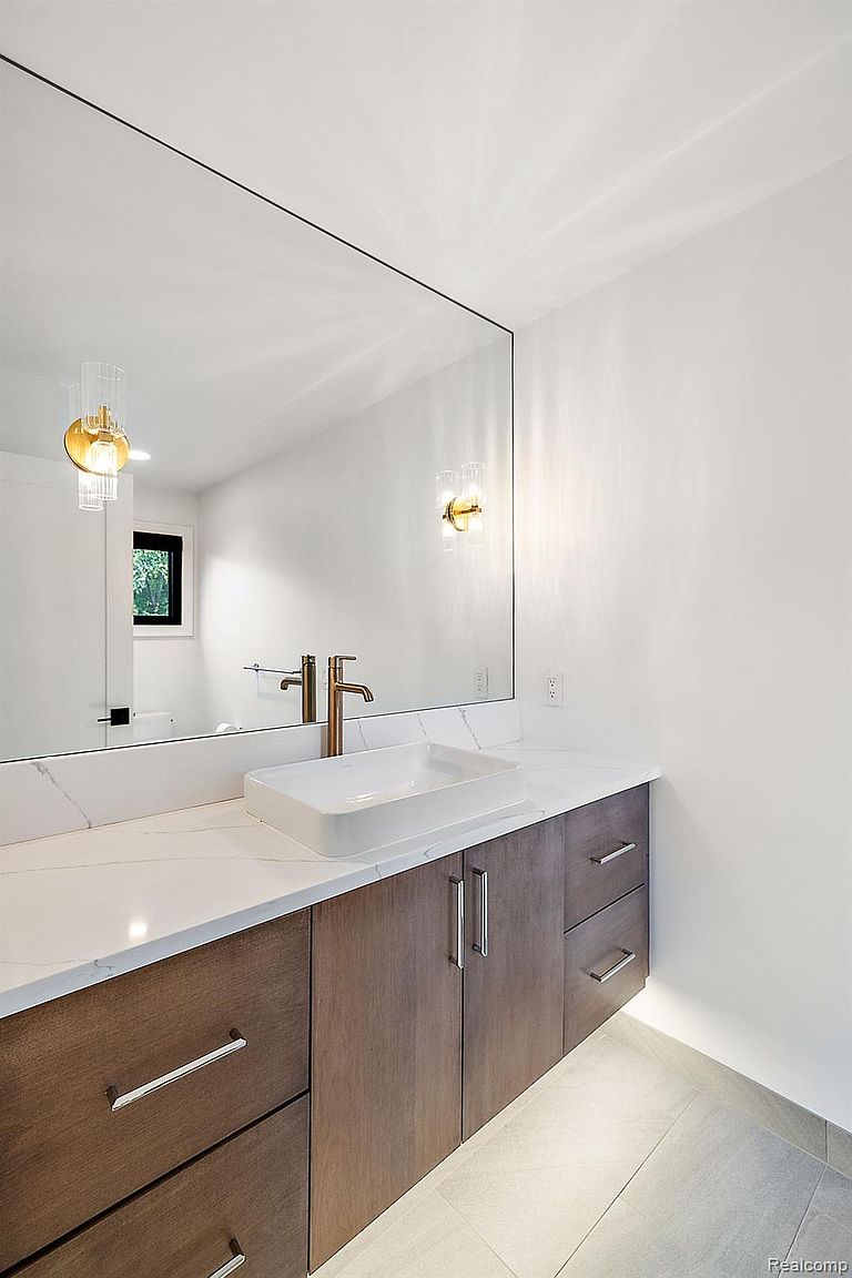 This is a modern bathroom featuring a sleek vanity with a white countertop and a rectangular vessel sink. The vanity has dark wood-tone cabinets with silver hardware, and a large frameless mirror hangs above. Gold-toned sconces flank the mirror, adding a touch of elegance, and the overall aesthetic is clean and contemporary.