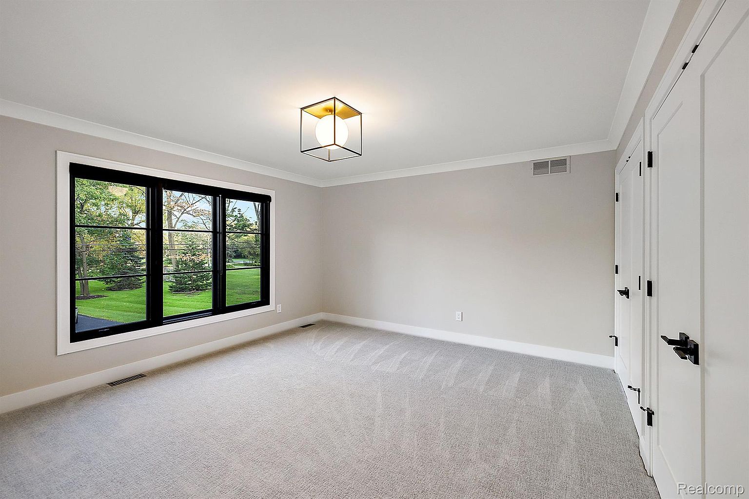 The image showcases a bright, empty bedroom with neutral-toned walls and light gray carpeting. A large window with black frames offers a view of a green lawn and trees, while a modern, cube-shaped light fixture illuminates the room. The space feels clean and spacious, ready for personalization.
