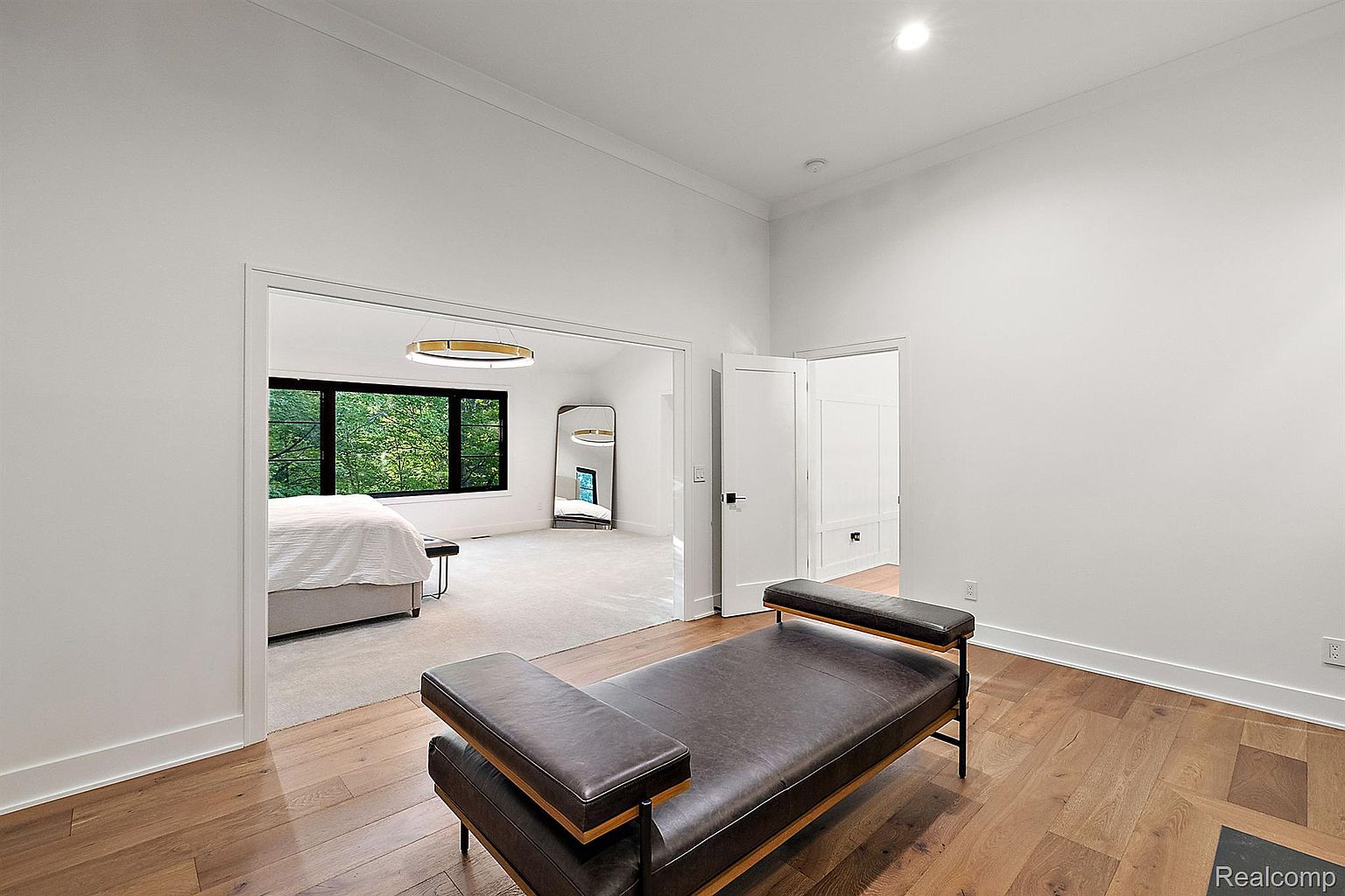This is a bright and spacious primary bedroom featuring hardwood floors and white walls. A modern leather bench sits in the foreground, leading the eye towards an open doorway to an adjacent room with a large window and a full-length mirror. The room exudes a clean and minimalist aesthetic.