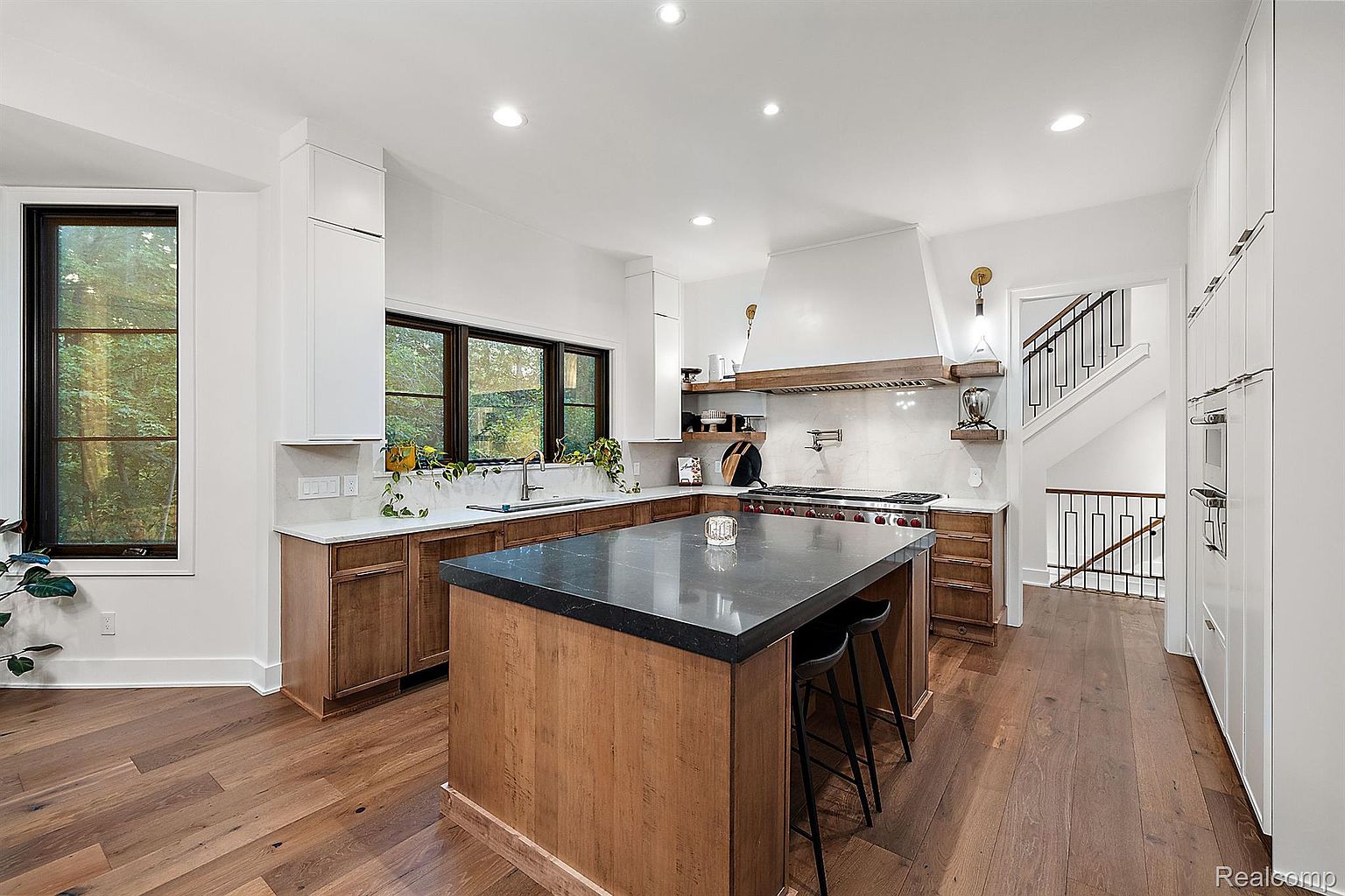 This is a bright and modern kitchen featuring custom wood cabinetry, a large center island with a dark countertop, and stainless steel appliances. The kitchen has a clean, contemporary design with a focus on natural light from the large windows. The hardwood floors add warmth to the space, and the open layout leads to a staircase, enhancing the home's flow.