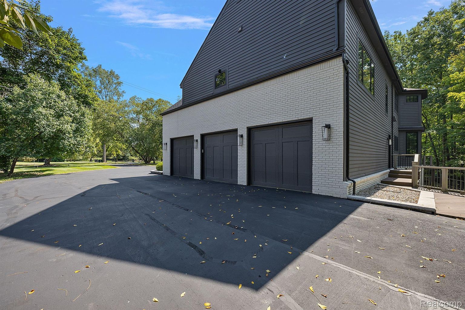 This image showcases a modern home's exterior, specifically the garage area. The garage features three dark-colored doors set against a light brick facade, with dark siding above. The driveway is paved and shows some fallen leaves, and the overall impression is clean and contemporary.