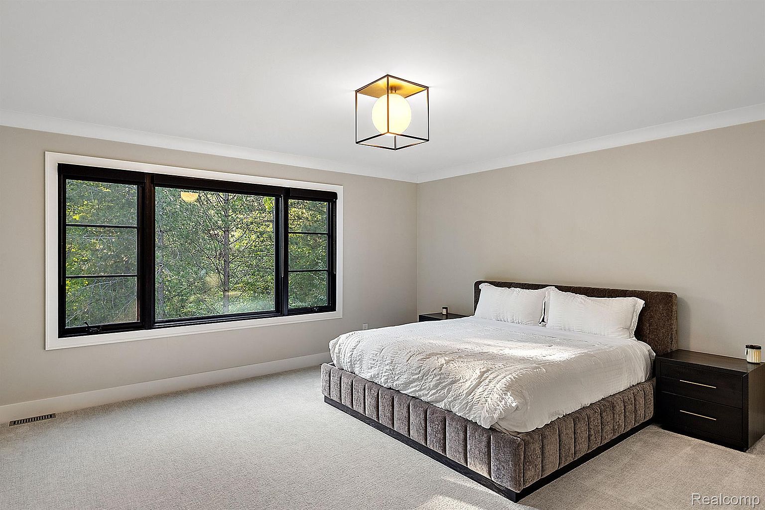 This is a primary bedroom featuring a large bed with a textured headboard and white bedding. A modern, cube-shaped light fixture hangs from the ceiling, and a large window provides natural light and a view of the outdoors. Two dark wood nightstands flank the bed, and the room is carpeted in a light neutral tone, creating a serene and inviting atmosphere.