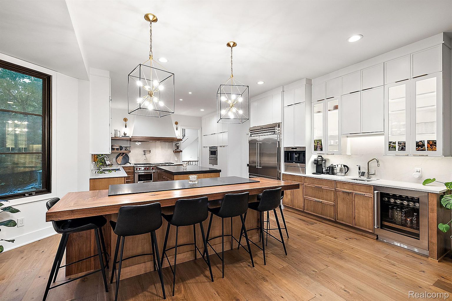 This is a bright and modern kitchen featuring a large wooden island with black countertops and seating for five. The kitchen boasts a combination of white and wood cabinetry, stainless steel appliances, and stylish pendant lighting. The hardwood flooring adds warmth to the space, creating an inviting atmosphere.