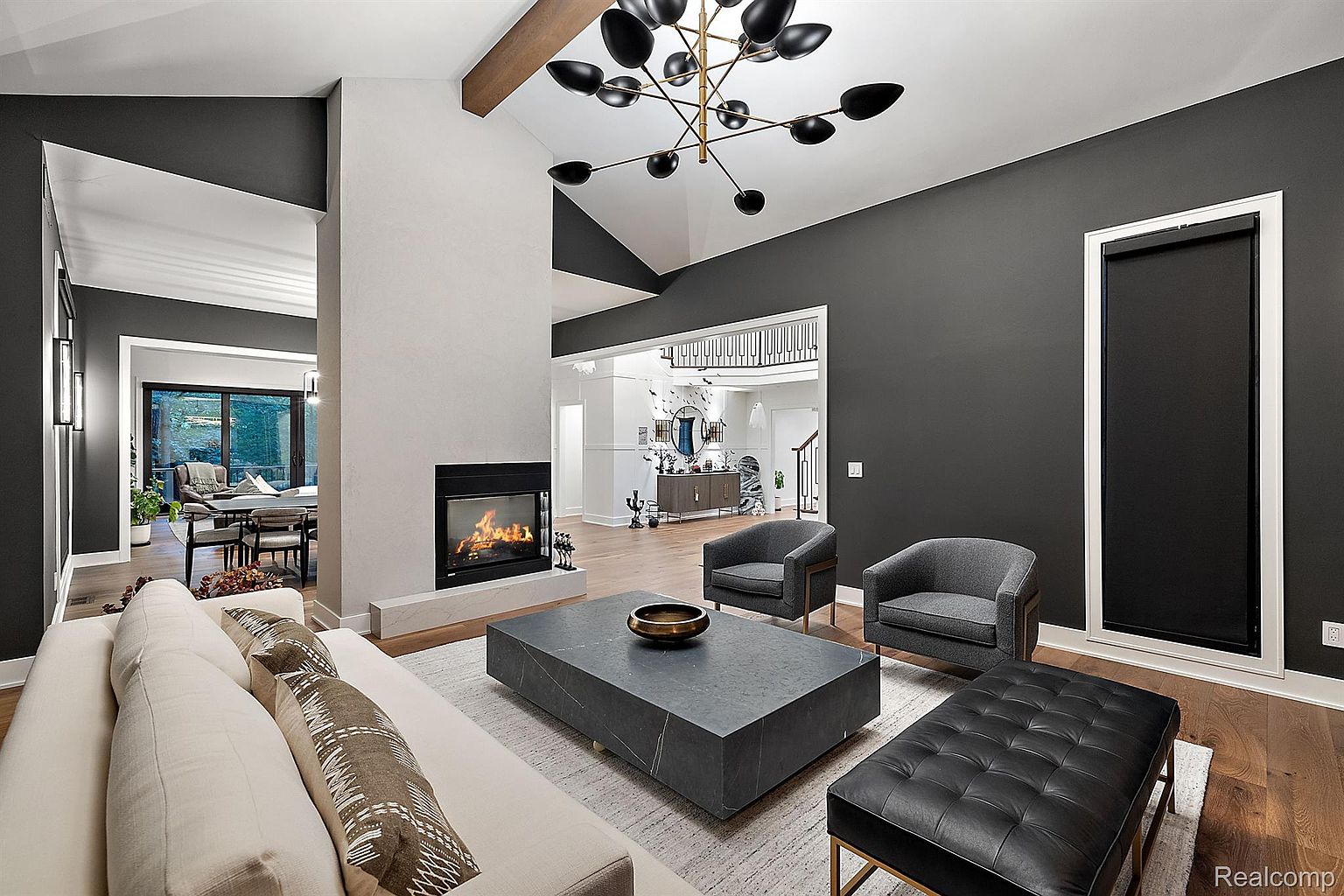 This is a stylish living room featuring a modern fireplace, a large gray coffee table, and comfortable seating. The room is decorated in a contemporary style with a neutral color palette and dark accent walls. A unique chandelier hangs from the ceiling, adding a touch of elegance.