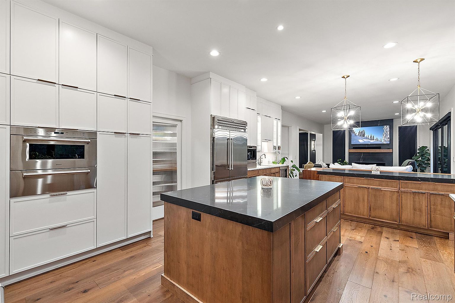 This is a bright and modern kitchen featuring white cabinetry, stainless steel appliances, and a large wooden island with a dark countertop. The open-concept design flows into a living area with a fireplace and television, creating a seamless living space. Hardwood floors add warmth and elegance to the space.