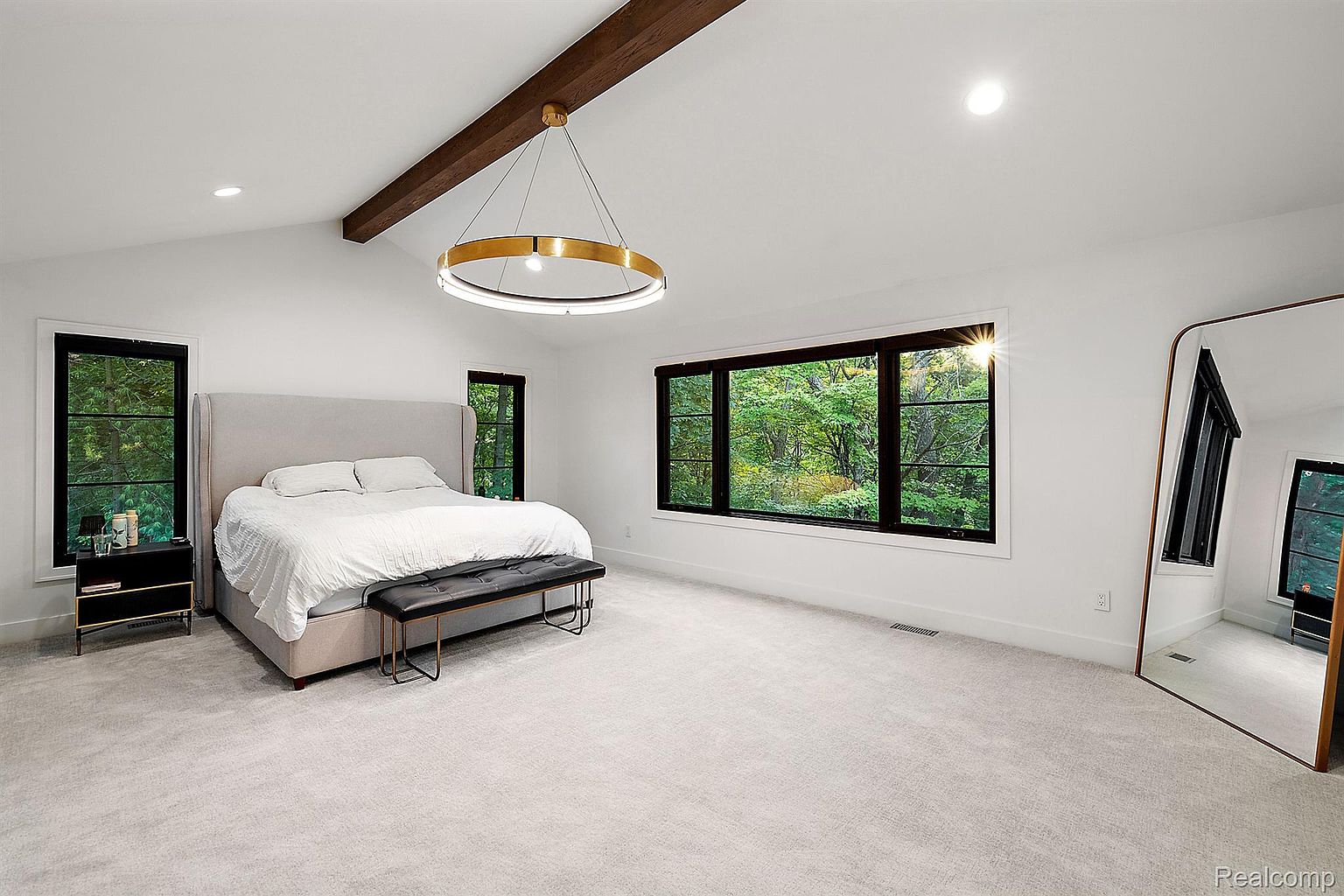This is a bright and spacious primary bedroom featuring a large bed with a light gray headboard and white bedding, complemented by a black leather bench. The room has white walls, light gray carpet, and black-framed windows offering natural light and views of the surrounding greenery. A modern, circular chandelier hangs from a wooden beam, adding a touch of elegance.