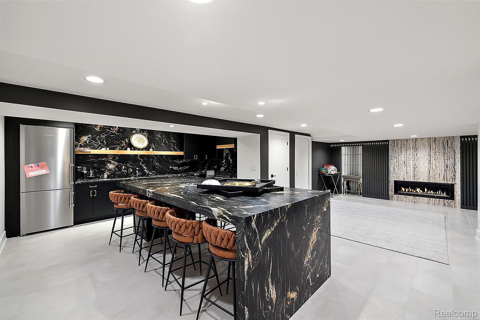 This is a modern kitchen featuring a large island with a striking black and gold marble countertop and seating for five. The kitchen includes stainless steel appliances, black cabinetry, and a unique marble backsplash. A fireplace adds a cozy touch to the open-concept space, which is well-lit with recessed lighting and has light-colored tile flooring.