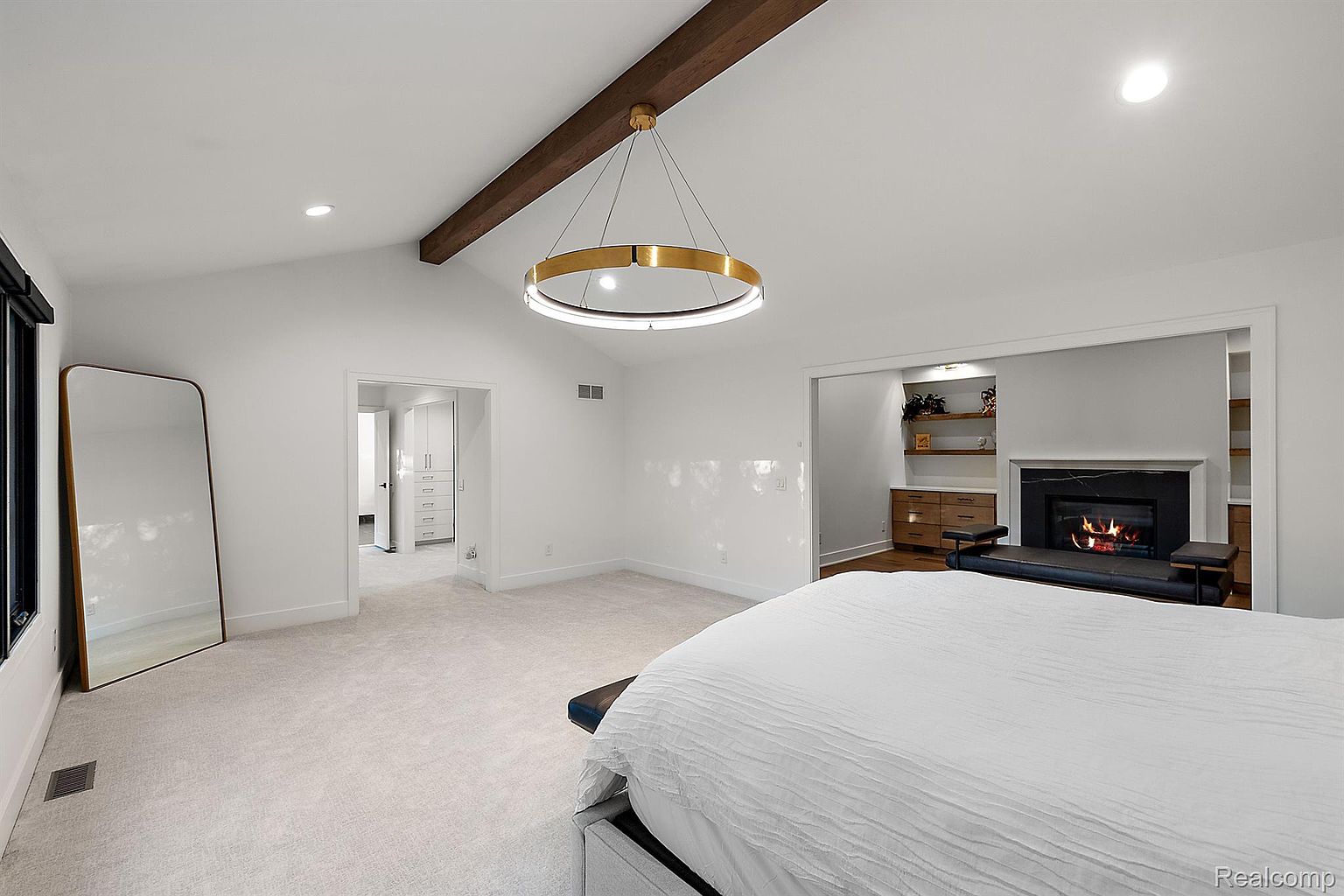 This is a bright and spacious primary bedroom featuring a large bed with white linens, a modern circular chandelier, and a cozy fireplace. The room has neutral-toned carpet, white walls, and a wooden beam on the ceiling, creating a serene and inviting atmosphere. A full-length mirror and doorways to other rooms add to the functionality of the space.