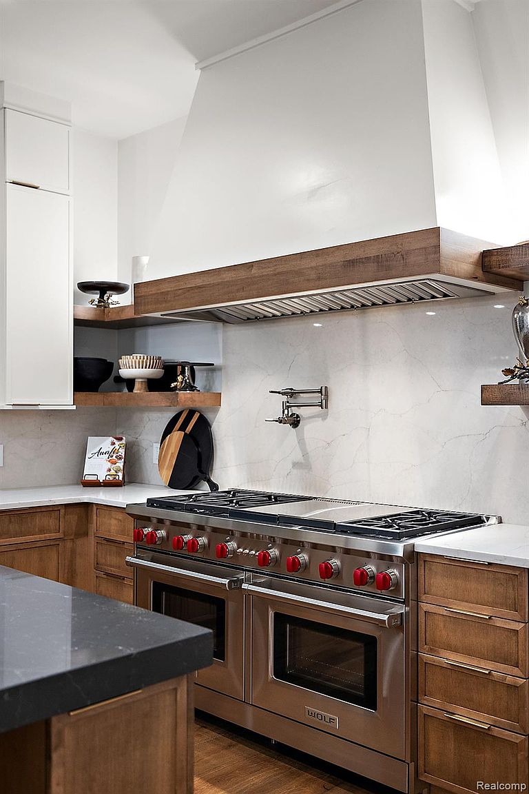 This is a detailed shot of a high-end kitchen featuring a professional-grade Wolf range with double ovens and red knobs. The range is set against a marble backsplash and beneath a custom wood-trimmed range hood. The cabinetry is a warm wood tone, and the countertops are a light, contrasting color, creating a luxurious and functional cooking space.