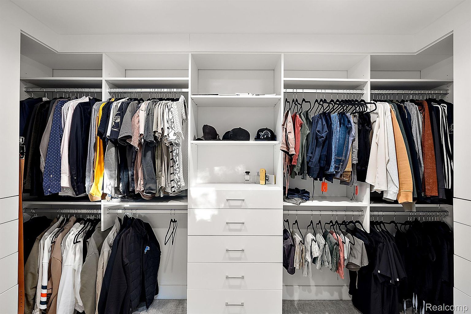 This is a well-organized walk-in closet featuring a white shelving system with hanging rods, drawers, and open shelves. Clothes are neatly arranged, and hats and accessories are displayed on the shelves. The closet appears spacious and designed for efficient storage.