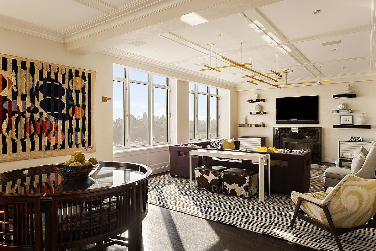 This sophisticated living room features a high-end, contemporary design with a dark wood circular table in the foreground and a plush, deep-toned sectional sofa arranged around a fireplace. The space is accented by a bold, abstract wall tapestry, a modern geometric area rug, and a striking gold-toned linear chandelier that draws the eye upward. Large windows provide an expansive, bright view of the city skyline, creating an airy and luxurious atmosphere.