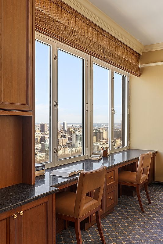 This sophisticated home office features a long, dark granite desk built directly beneath a series of large windows that offer a stunning, elevated cityscape view. The space is furnished with two matching leather chairs and rich wood cabinetry, creating a professional and refined atmosphere. The combination of natural light, high-rise views, and custom millwork makes this an ideal workspace for productivity and inspiration.