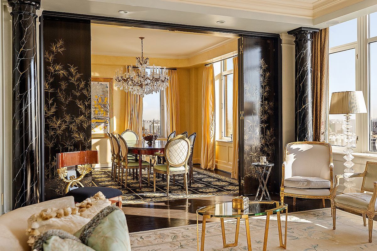 This sophisticated living space features a grand, open-plan layout that seamlessly transitions into a formal dining room through ornate, black-lacquered folding doors adorned with gold bamboo motifs. The foreground showcases elegant cream-colored seating and a glass-topped coffee table, while the background highlights a luxurious dining area illuminated by a sparkling crystal chandelier. The room is framed by dramatic black marble columns, creating a high-end, opulent atmosphere with a view of the city skyline through large windows.