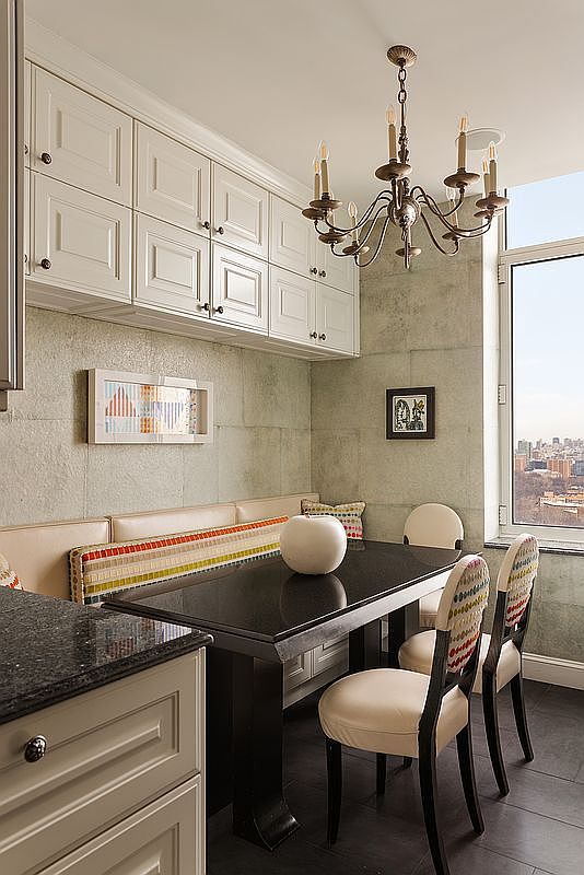 This elegant dining nook features a built-in banquette with colorful, patterned upholstery paired with a sleek, dark granite-topped table and two matching chairs. The space is illuminated by a classic chandelier and framed by white cabinetry above the seating area, while a large window provides a stunning urban view. The combination of textured walls and sophisticated furniture creates a cozy yet refined atmosphere perfect for intimate meals.