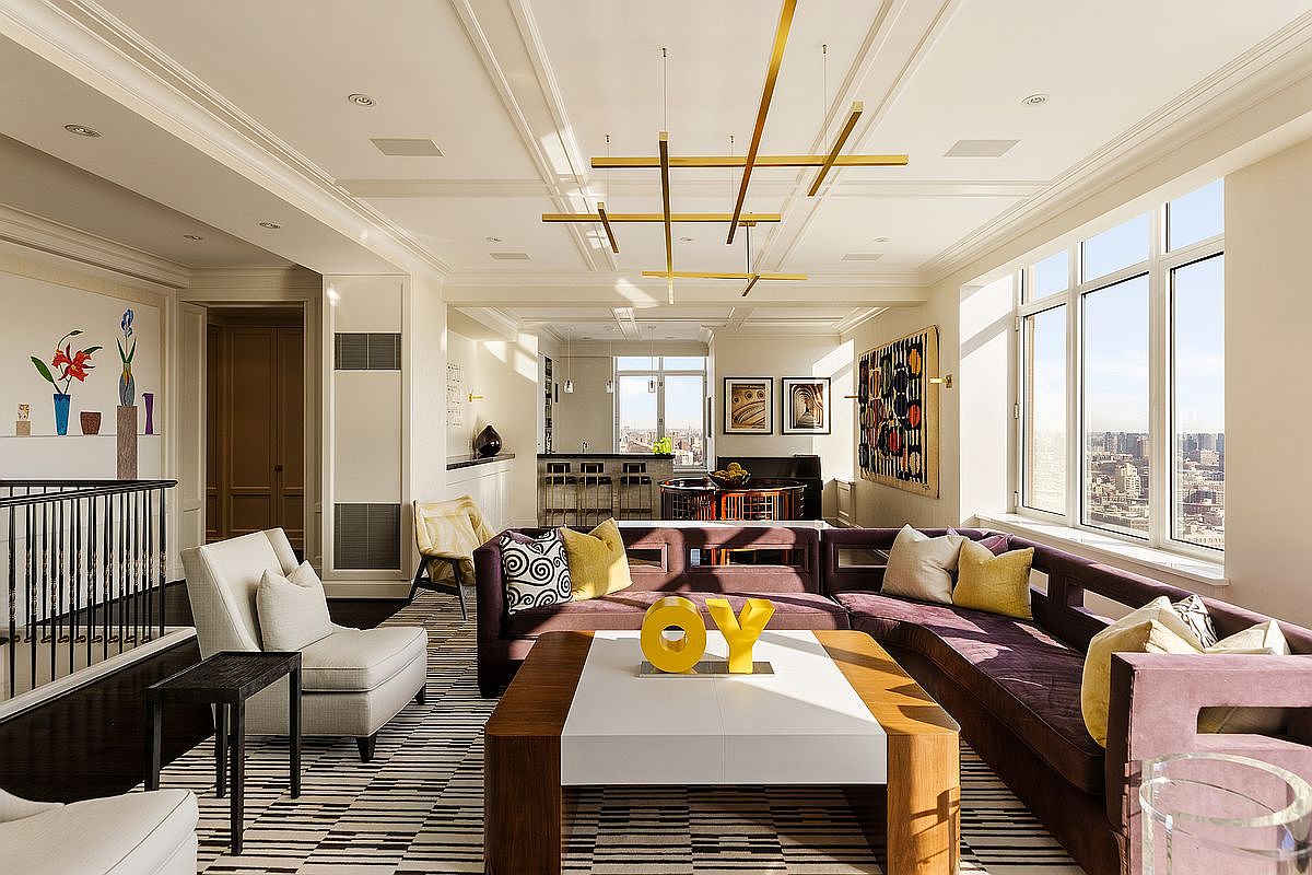 This sophisticated, high-rise living room features a spacious open-concept layout with a large, plush purple sectional sofa and a modern white-topped coffee table adorned with a bold yellow 'OY' sculpture. The room is illuminated by a striking, geometric gold chandelier and benefits from abundant natural light streaming through expansive windows that offer panoramic city views. The interior design blends contemporary luxury with artistic flair, highlighted by a vibrant wall tapestry and a sleek, striped area rug.