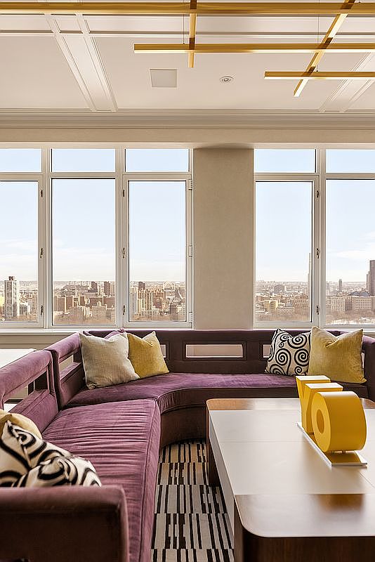 This sophisticated living room features a plush, curved purple velvet sectional sofa accented with patterned and solid yellow throw pillows. A large, modern coffee table sits in the foreground, while floor-to-ceiling windows provide a panoramic, high-rise view of the city skyline. The space is illuminated by a unique, geometric gold light fixture suspended from a coffered ceiling, creating an elegant and luxurious atmosphere.
