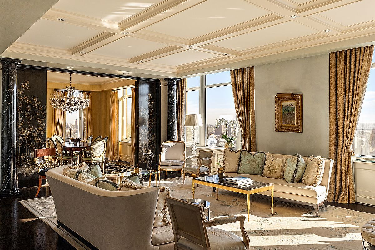 This elegant, high-end living room features a sophisticated blend of classic and contemporary design, highlighted by a coffered ceiling and expansive windows offering city views. The space is furnished with plush, neutral-toned seating, a sleek coffee table, and ornate decorative accents, all anchored by a large, patterned area rug. The perspective captures the open-concept flow into an adjacent dining area, emphasizing the luxurious and airy atmosphere of the residence.