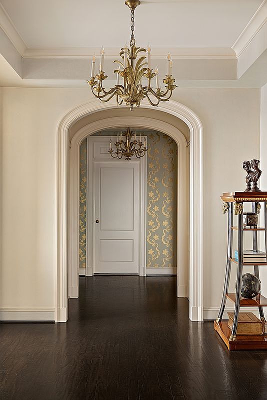 This elegant hallway features a dramatic arched entryway leading into a secondary space adorned with sophisticated gold-patterned wallpaper. The foreground is anchored by a dark hardwood floor and a classic, ornate gold chandelier, while a decorative tiered display stand adds a touch of traditional luxury to the right. The perspective is centered and symmetrical, creating a formal and inviting transition between rooms.