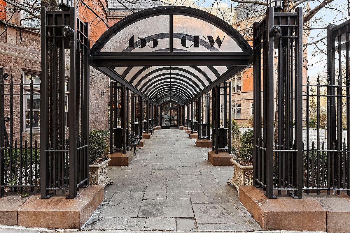 This image captures the grand, arched entryway of 455 CPW, featuring a striking black metal pergola that creates a covered walkway leading to the building's entrance. The path is paved with large stone slabs and flanked by decorative planters and ornate iron fencing, evoking a sense of historic elegance and exclusivity. The perspective is a centered, eye-level shot that draws the viewer's gaze directly toward the building's main door, emphasizing the sophisticated architectural character of the property.